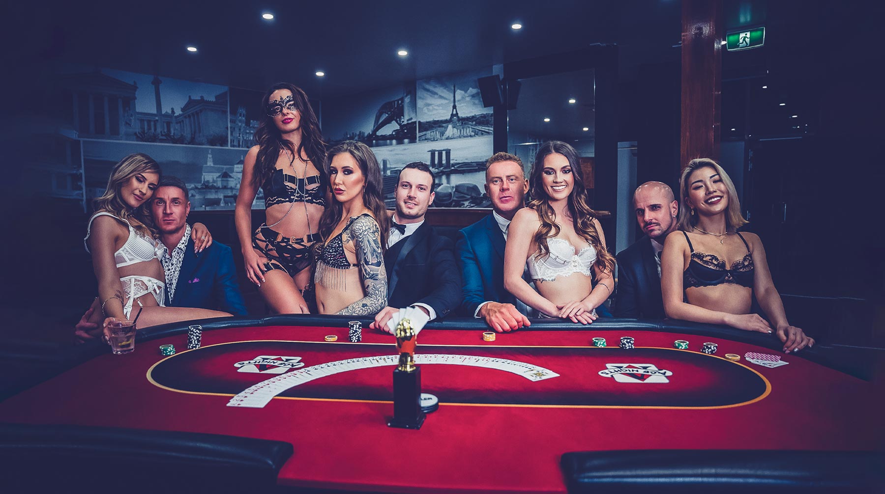 A group of elegantly dressed men and women, some in formal wear and others in lingerie, seated around a red blackjack table in a glamorous strip blackjack casino environment.