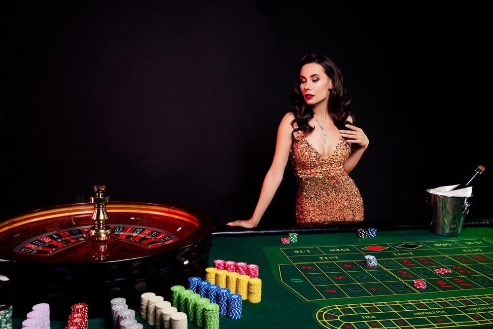 A woman in a glittering gold evening dress standing beside a live porn Roulette table with colorful chips, set against a dark background with a champagne bucket nearby.